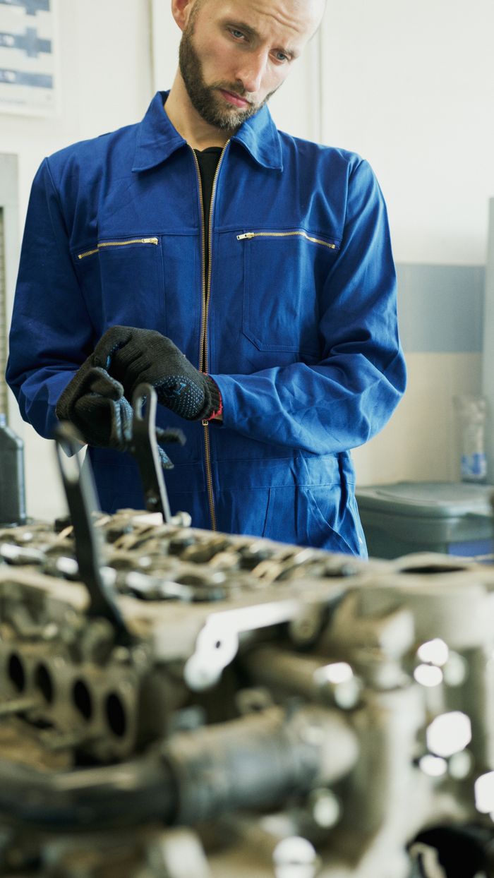 Mechanic wearing blue coveralls and gloves inspecting an engine in a workshop.