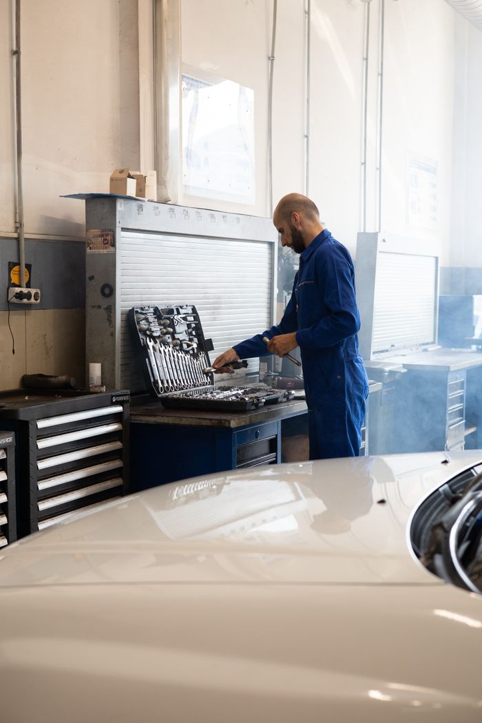 Mechanic in blue coveralls working in an automotive repair shop with tools and car in view.